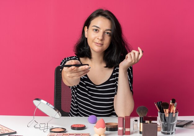 Pleased looking  young beautiful girl sits at table with makeup tools holding out eyeliner  isolated on pink wall