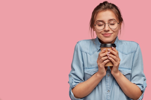 Piacevole bella donna caucasica tiene bevanda aromatica, beve cappuccino o caffè, si sente caldo, chiude gli occhi dal piacere