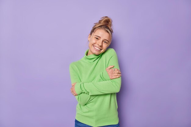 Pleased lovely woman with combed hair embraces herself feels comfortable wears green jumper smiles gently being self egoist poses against purple background Self love and acceptance concept