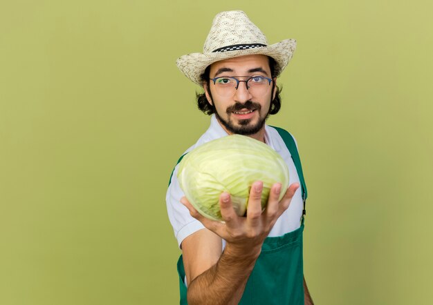 Pleased male gardener in optical glasses wearing gardening hat holds cabbage looking  