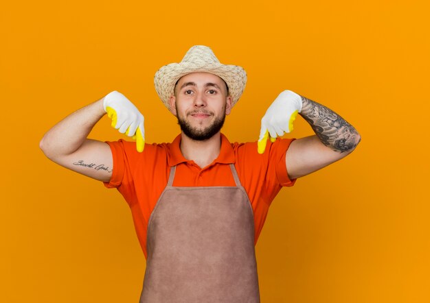 Pleased male gardener wearing gardening hat and gloves points down with two hands 