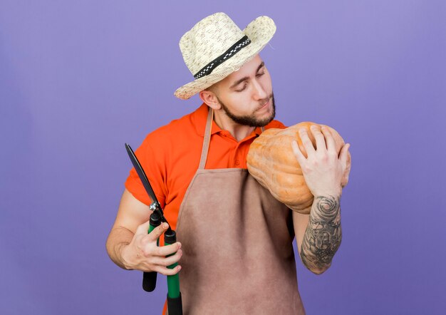 Pleased male gardener wearing gardening hat looks at pumpkin and holds clippers