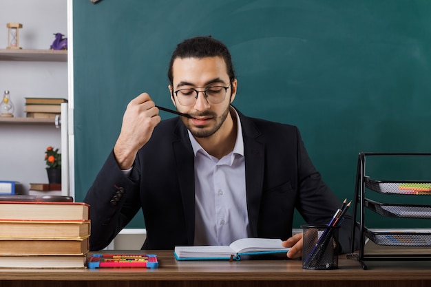 Pleased male teacher wearing glasses reading book putting pencil in mouth sitting at table with school tools in classroom