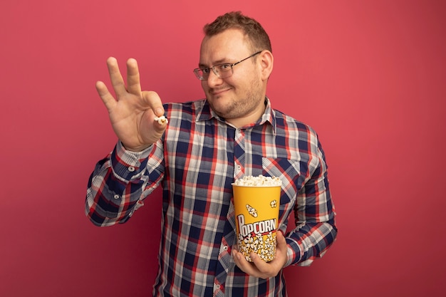 Pleased man in glasses and checked shirt holding bucket with popcorn smiling with happy face standing over pink wall