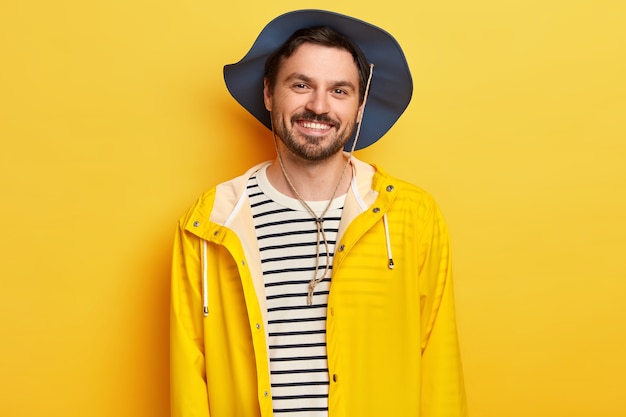 Pleased man wears hat, yellow raincoat, spends leisure time actively, poses indoor, expresses good emotions, has stubble