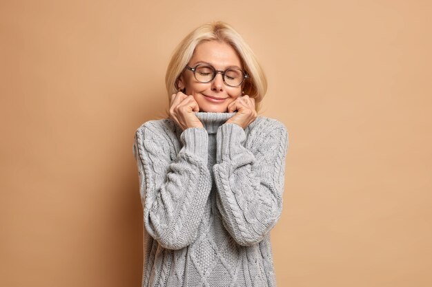 Pleased middle aged beautiful woman keeps hands on collar of warm sweater stands with closed eyes wears glasses recalls something pleasant.