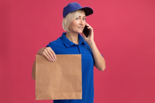 Pleased middle-aged blonde delivery woman in blue uniform and cap talking on phone holding paper package looking at front isolated on pink wall with copy space