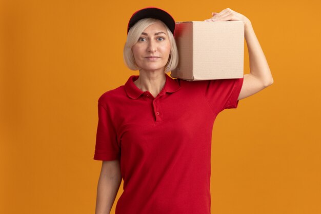 Pleased middle-aged blonde delivery woman in red uniform and cap holding cardboard box on shoulder looking at front isolated on orange wall with copy space