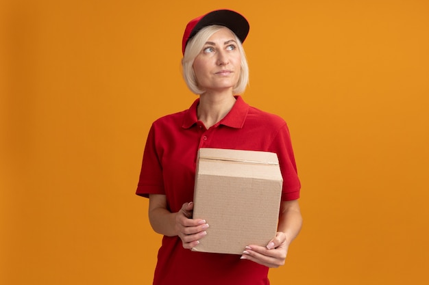 Pleased middle-aged blonde delivery woman in red uniform and cap holding cardbox looking up 
