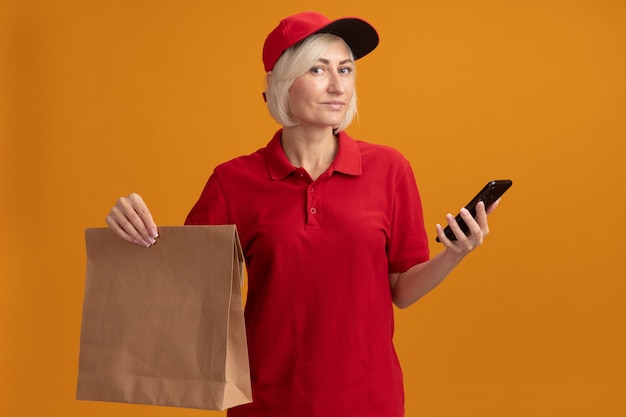 Pleased middle-aged blonde delivery woman in red uniform and cap holding paper package and mobile phone 