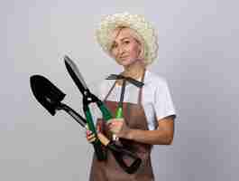 Free photo pleased middle-aged blonde gardener woman in uniform wearing hat looking at front holding gardener tools isolated on white wall with copy space