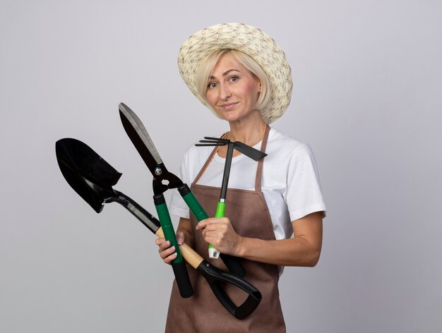 Pleased middle-aged blonde gardener woman in uniform wearing hat looking at front holding gardener tools isolated on white wall with copy space