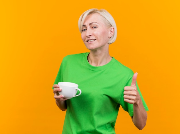 Pleased middle-aged blonde slavic woman holding cup of tea looking at camera showing thumb up isolated on yellow background with copy space