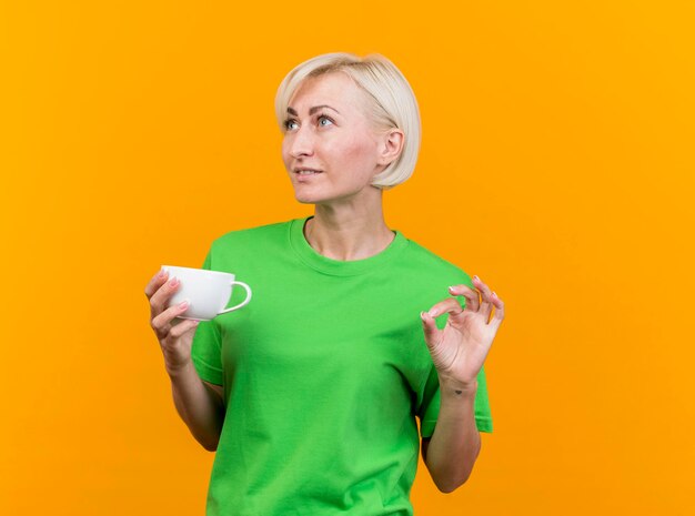 Pleased middle-aged blonde slavic woman holding cup of tea looking at side doing ok sign isolated on yellow wall with copy space