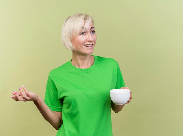 Pleased middle-aged blonde slavic woman holding cup of tea looking at side showing empty hand isolated on olive green wall with copy space