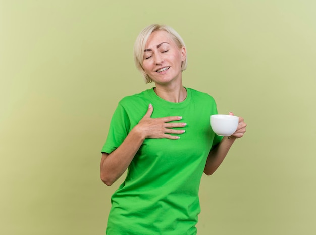 Pleased middle-aged blonde slavic woman holding cup of tea putting hand on chest with closed eyes isolated on olive green wall with copy space