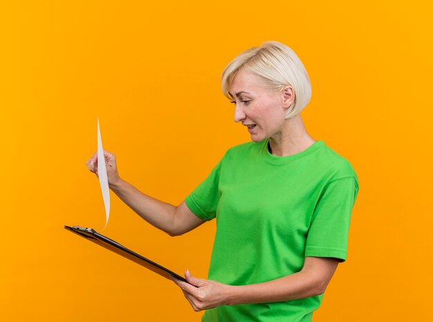 Pleased middle-aged blonde slavic woman holding and looking at clipboard isolated on yellow background