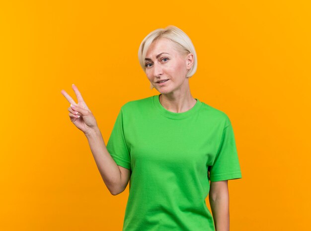 Pleased middle-aged blonde slavic woman looking at camera doing peace sign isolated on yellow background with copy space