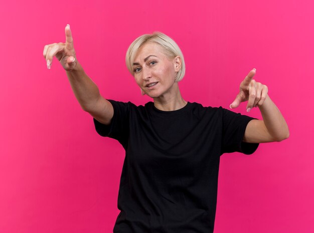 Pleased middle-aged blonde slavic woman looking at camera pointing straight isolated on crimson background