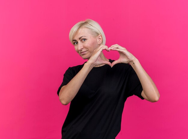 Довольная белокурая славянская женщина средних лет, смотрящая вперед, делает знак сердца, изолированную на розовой стене с копией пространства