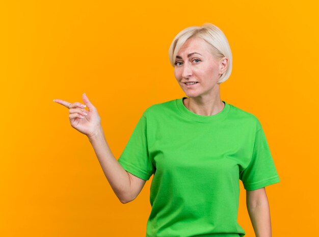 Pleased middle-aged blonde slavic woman looking at front pointing at side isolated on yellow wall