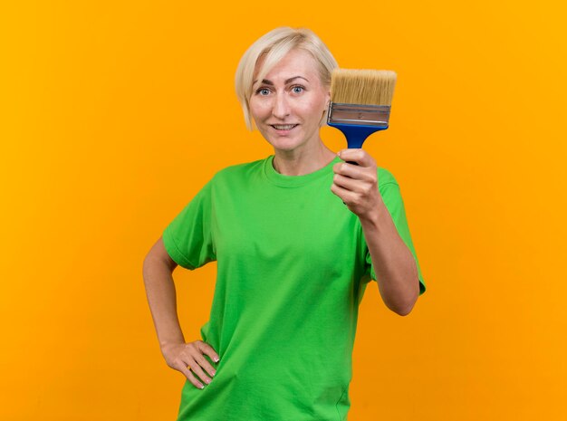 Pleased middle-aged blonde slavic woman looking at front showing paint brush keeping hand on waist isolated on yellow wall with copy space