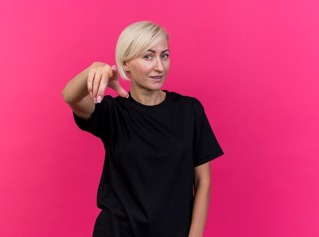 Free photo pleased middle-aged blonde slavic woman looking and pointing at camera isolated on crimson background with copy space