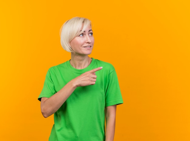 Pleased middle-aged blonde slavic woman looking and pointing at side isolated on yellow background with copy space