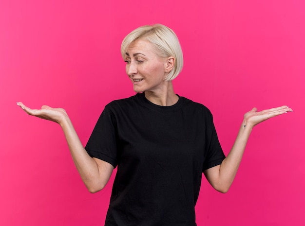 Pleased middle-aged blonde slavic woman showing empty hands looking at one of them isolated on pink wall