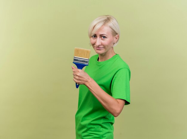 Pleased middle-aged blonde slavic woman standing in profile view  holding paint brush isolated on olive green wall with copy space