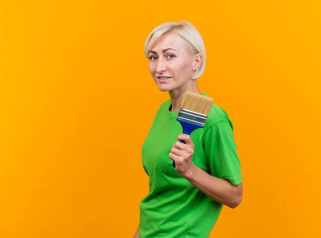 Pleased middle-aged blonde slavic woman standing in profile view  holding paint brush isolated on yellow wall with copy space