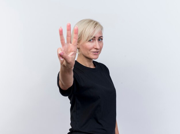 Pleased middle-aged blonde slavic woman standing in profile view looking at camera showing three with hand isolated on white background with copy space