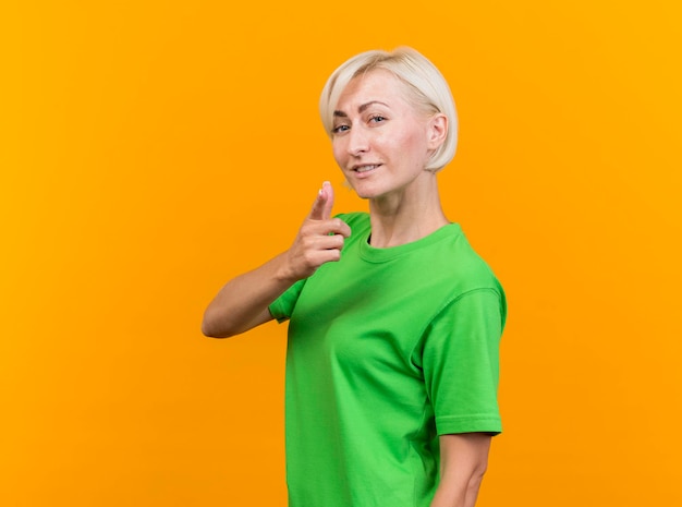Pleased middle-aged blonde slavic woman standing in profile view looking at front doing pistol gesture isolated on yellow wall with copy space