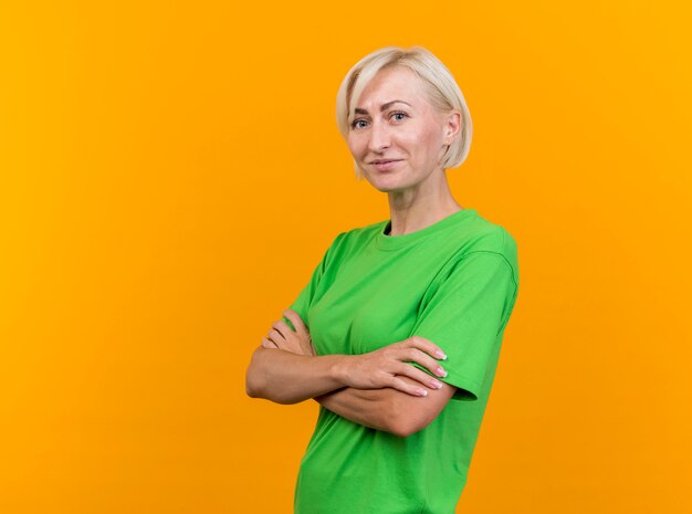 Pleased middle-aged blonde slavic woman standing with closed posture in profile view looking at camera isolated on yellow background with copy space