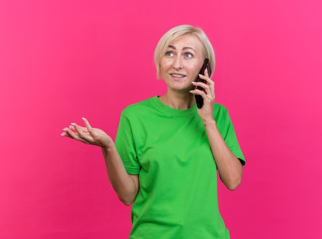 Pleased middle-aged blonde slavic woman talking on phone showing empty hand looking at side isolated on crimson background with copy space