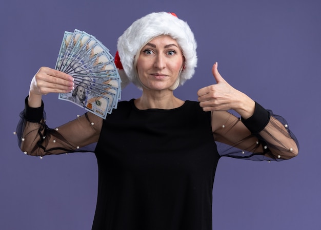 Lieta donna bionda di mezza età che indossa il cappello di natale tenendo i soldi guardando la telecamera che mostra il pollice in alto isolato su sfondo viola