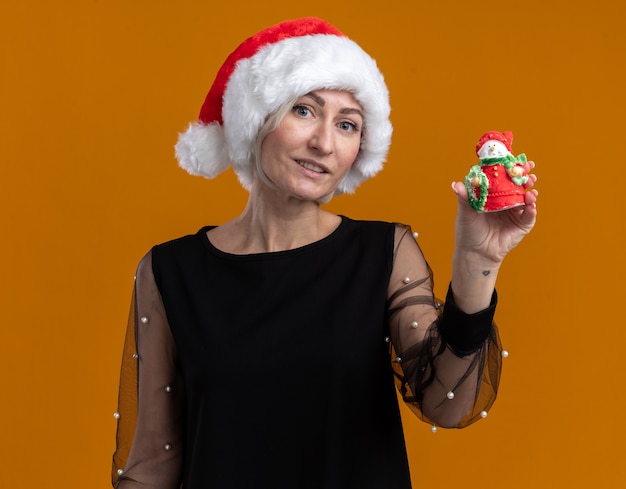 Pleased middle-aged blonde woman wearing christmas hat looking at camera showing small snowman statue to camera isolated on orange background