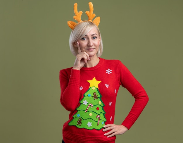 Foto gratuita compiaciuta donna bionda di mezza età che indossa la fascia di corna di renna di natale e maglione di natale cercando di tenere la mano sulla vita e sul mento isolato sulla parete verde oliva con spazio di copia