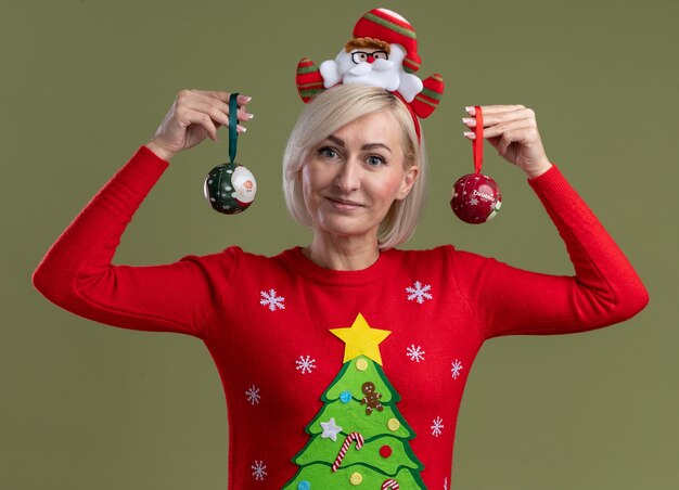 Pleased middle-aged blonde woman wearing santa claus headband and christmas sweater holding christmas baubles near head looking at camera isolated on olive green background