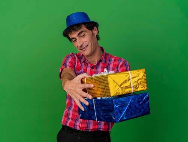 Довольный кавказский тусовщик средних лет в партийной шляпе протягивает подарочные пакеты к камере и смотрит на них, изолированные на зеленом фоне с копией пространства