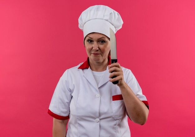  pleased middle-aged female cook in chef uniform holding knife on isolated pink wall