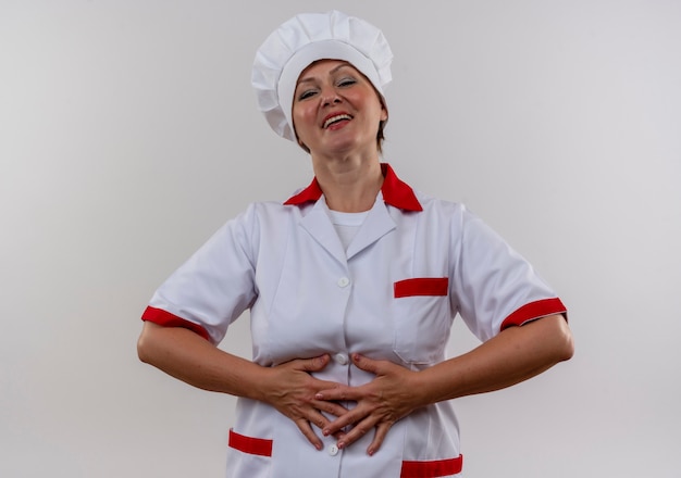 Довольная женщина-повар средних лет в униформе шеф-повара кладет руку на живот на изолированной белой стене
