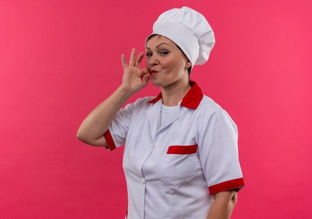 Pleased middle-aged female cook in chef uniform showing delicious gesture on isolated pink wall with copy space