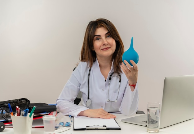 Pleased middle-aged female doctor wearing medical robe and stethoscope sitting at desk with medical tools clipboard and laptop holding enema isolated