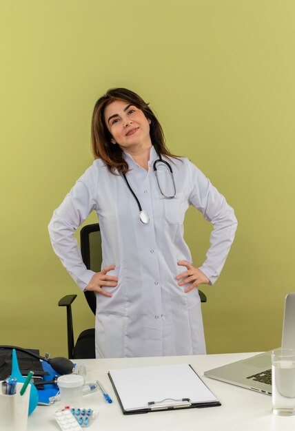 Pleased middle-aged female doctor wearing medical robe and stethoscope standing behind desk with medical tools clipboard and laptop keeping hands on waist isolated