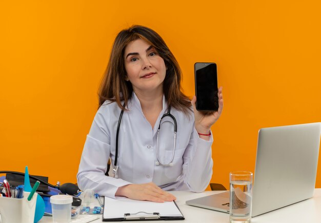 청진 기 책상에 앉아 의료 가운을 입고 기쁘게 중년 여성 의사 복사 공간이 격리 된 주황색 벽에 전화를 들고 의료 도구와 노트북에서 작동