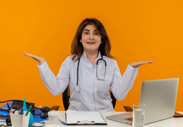 Pleased middle-aged female doctor wearing medical robe with stethoscope sitting at desk work on laptop with medical tools spreads hands on isolated orange wall with copy space