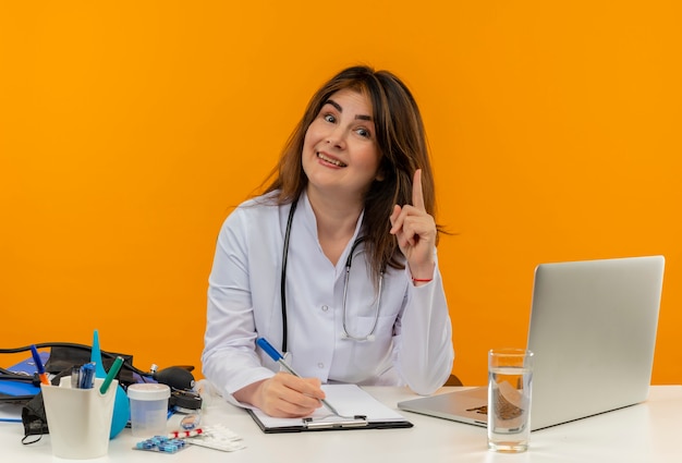 Довольная женщина-врач средних лет в медицинском халате со стетоскопом, сидя за столом, работает на ноутбуке с медицинскими инструментами, пишет в буфере обмена указывает на изолированную оранжевую стену с копией пространства