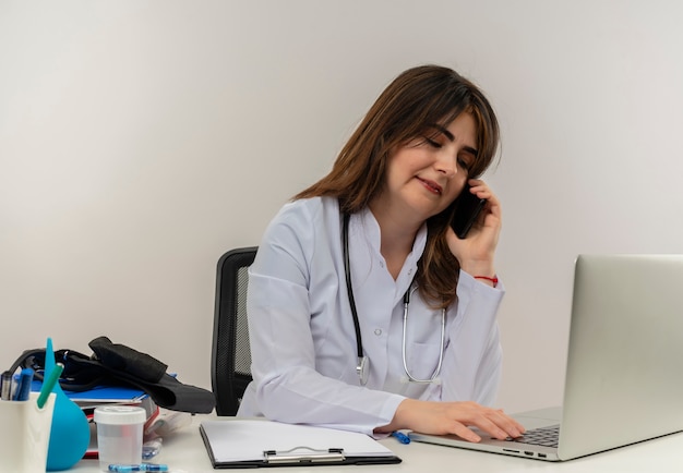 청진 기 의료 도구와 노트북에 책상 작업에 앉아 의료 가운을 입고 기쁘게 중년 여성 의사는 전화로 말하고 흰 벽에 노트북을 사용