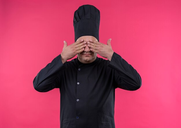 Pleased middle-aged male cook in chef uniform covered eyes with hands on isolated pink wall